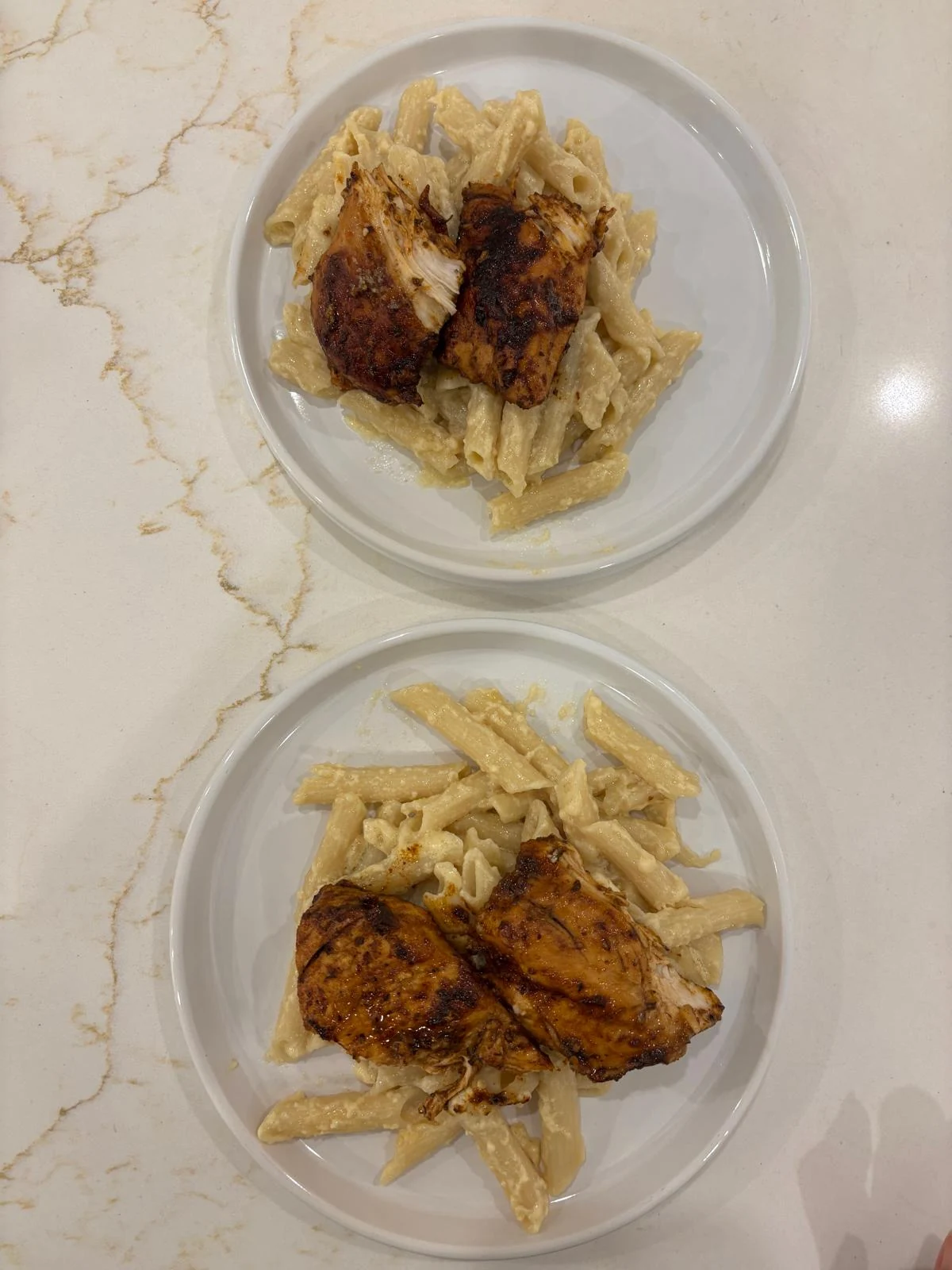 Two plates of creamy penne topped with browned seasoned fillet pieces. Exposed interiors have long white chicken-breast-like fibers.