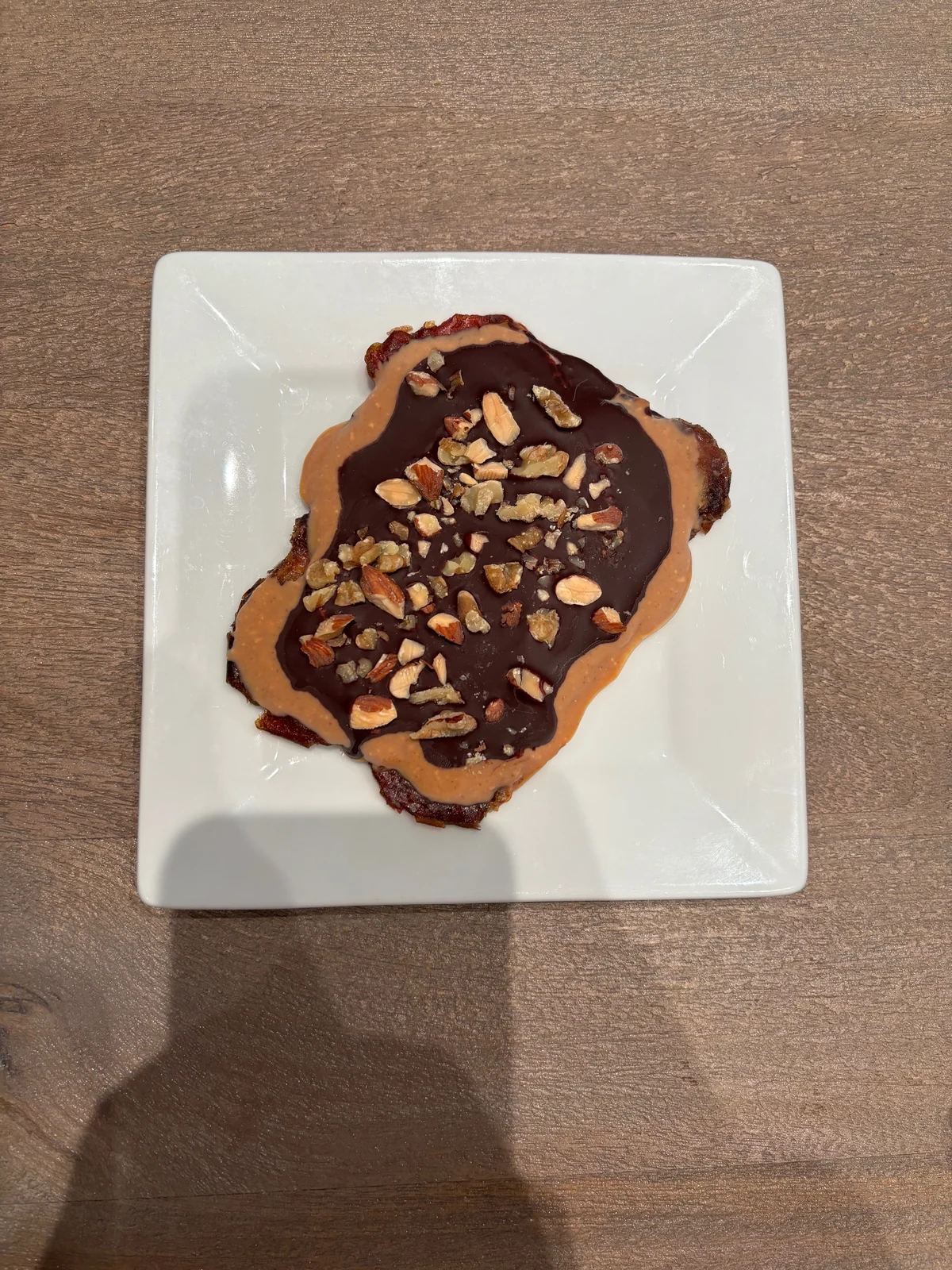 Thin irregular dark reddish-brown base with tan nut butter, glossy dark chocolate and chopped nuts. Exposed base edges resemble pressed dates. Chopped almond and walnut shapes are visible.
