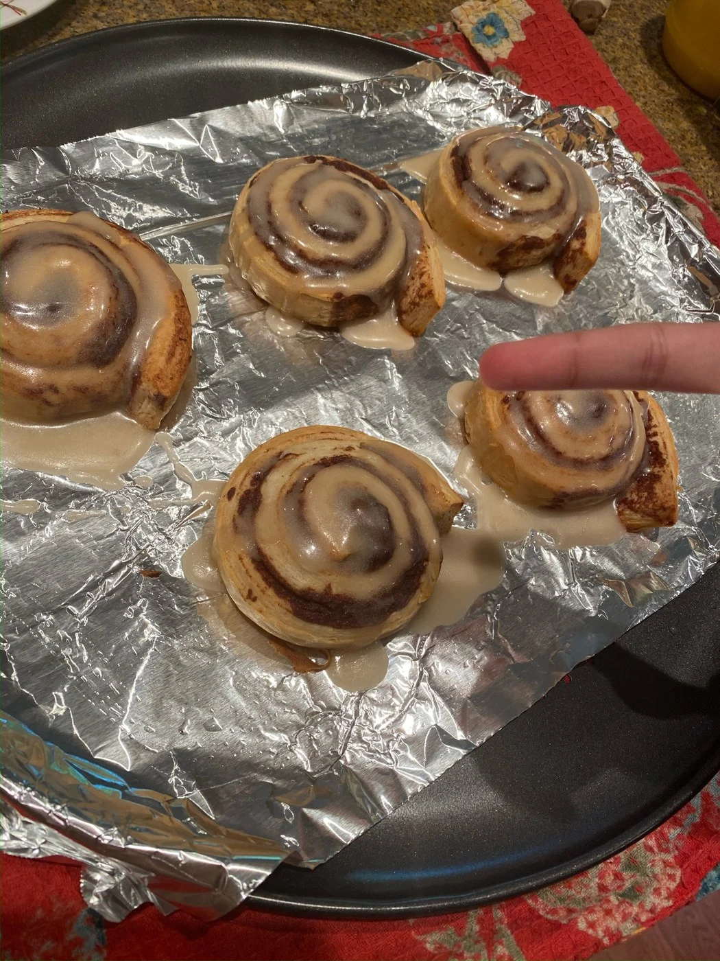 A finger pointing toward a glazed cinnamon swirl on a foil-lined tray.
