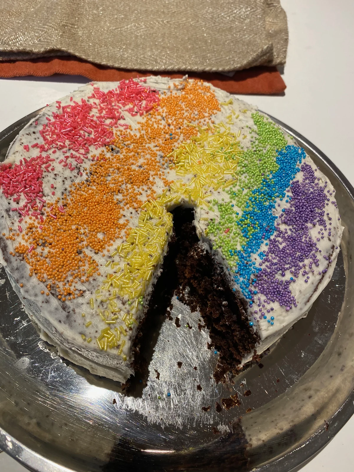 Pale exterior frosting, rainbow stripes of sprinkles and a wedge removed revealing dark cake crumb.