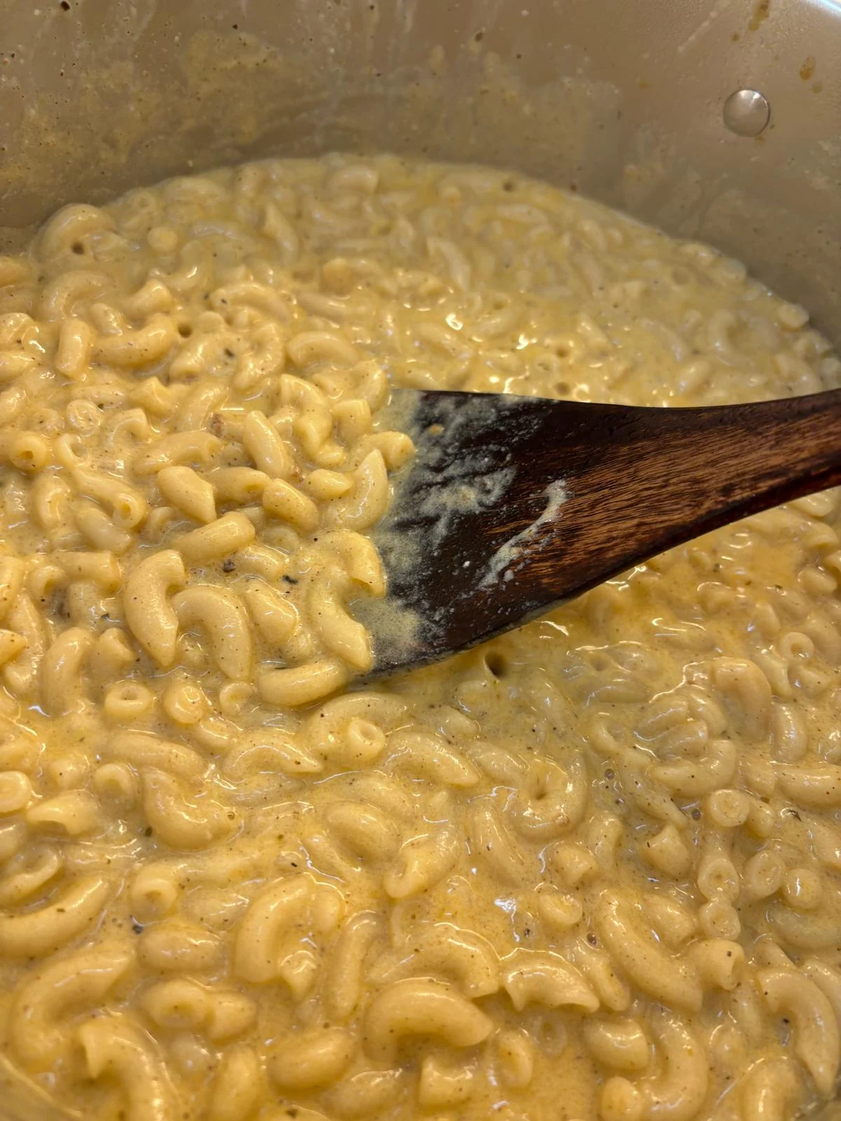 Elbow macaroni in abundant pale yellow cheese-like sauce in a pot, stirred with wooden spoon.